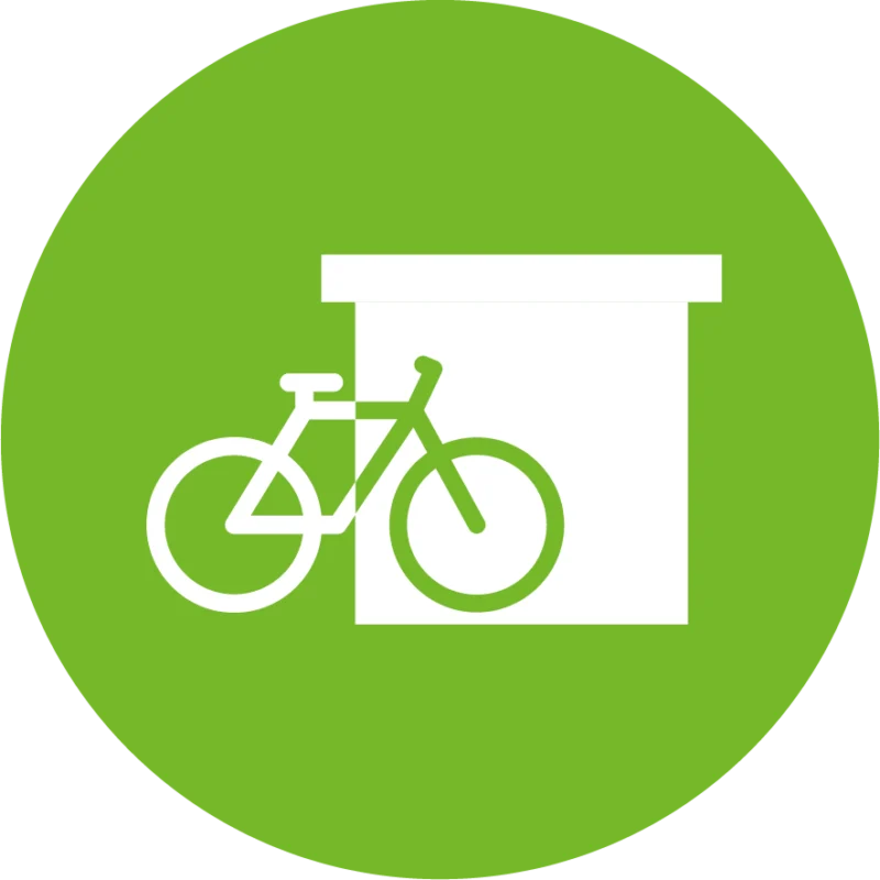 Weißes Piktogramm eines Fahrrads vor einem geschlossenen Abstellraum auf grünem Kreis – symbolisiert einen sicheren Fahrradstellplatz.